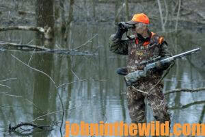 A hunter stands by a stream and scouts out the area through his binoculars